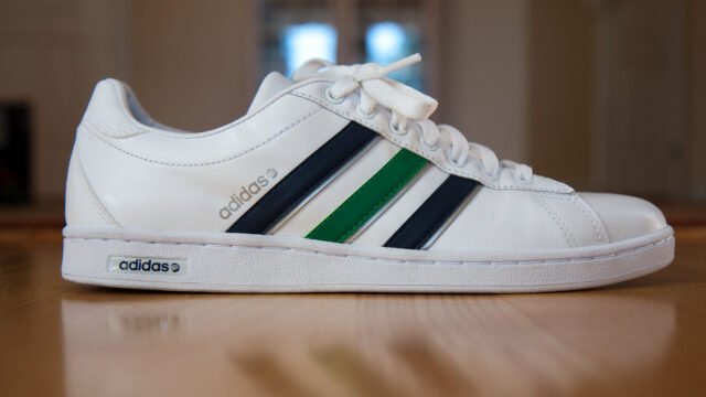 Бяла кожена маратонка Adidas с тъмносини и зелена ивица върху дървен под