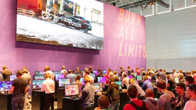 Посетители играят Need for Speed Heat на щанда на играта на Gamescom 2019, а на големия екран се вижда състезание