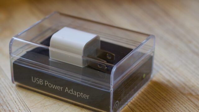 Малко бяло USB зарядно на Apple в прозрачна пластмасова опаковка върху дървена маса