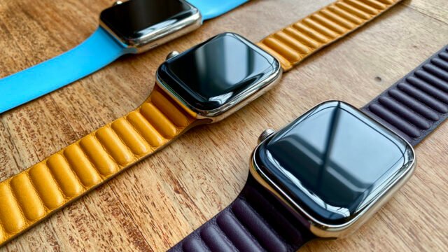 Три часовника Apple Watch от различни поколения с цветни каишки върху дървена маса