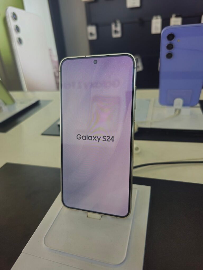 Смартфон Samsung Galaxy S24 с надпис Galaxy S24 на екрана, поставен на стойка в магазин