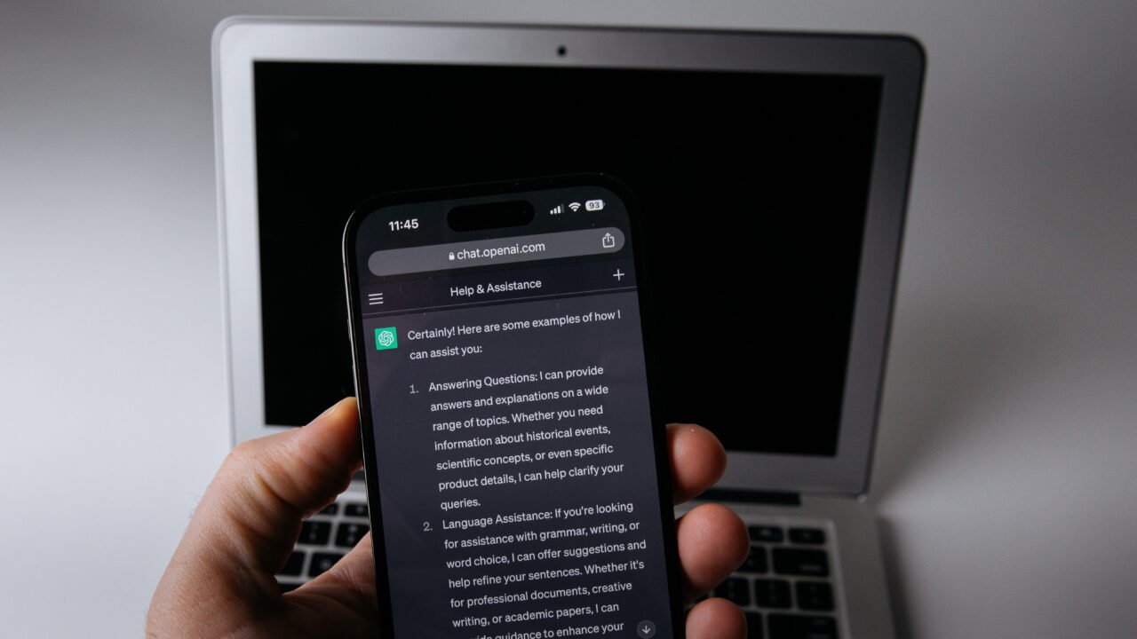 Ръка държи смартфон с отворен чатбот пред лаптоп – shadow AI и рискът от изтичане на фирмени данни