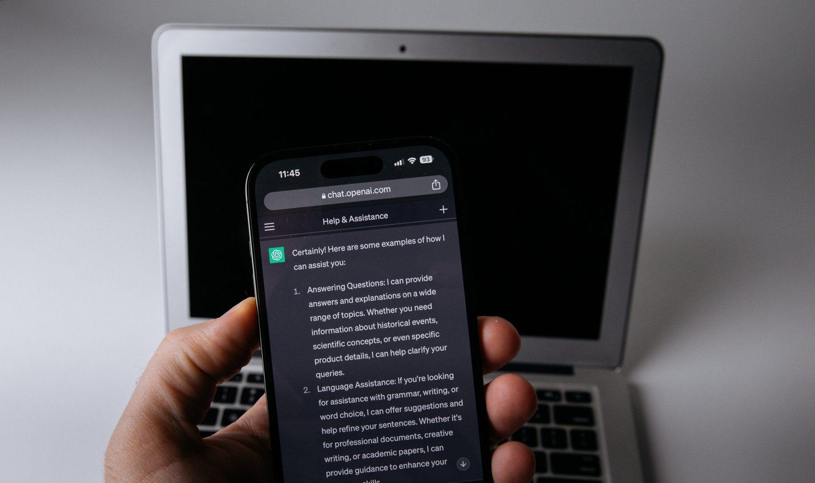 Ръка държи смартфон с отворен чатбот пред лаптоп – shadow AI и рискът от изтичане на фирмени данни